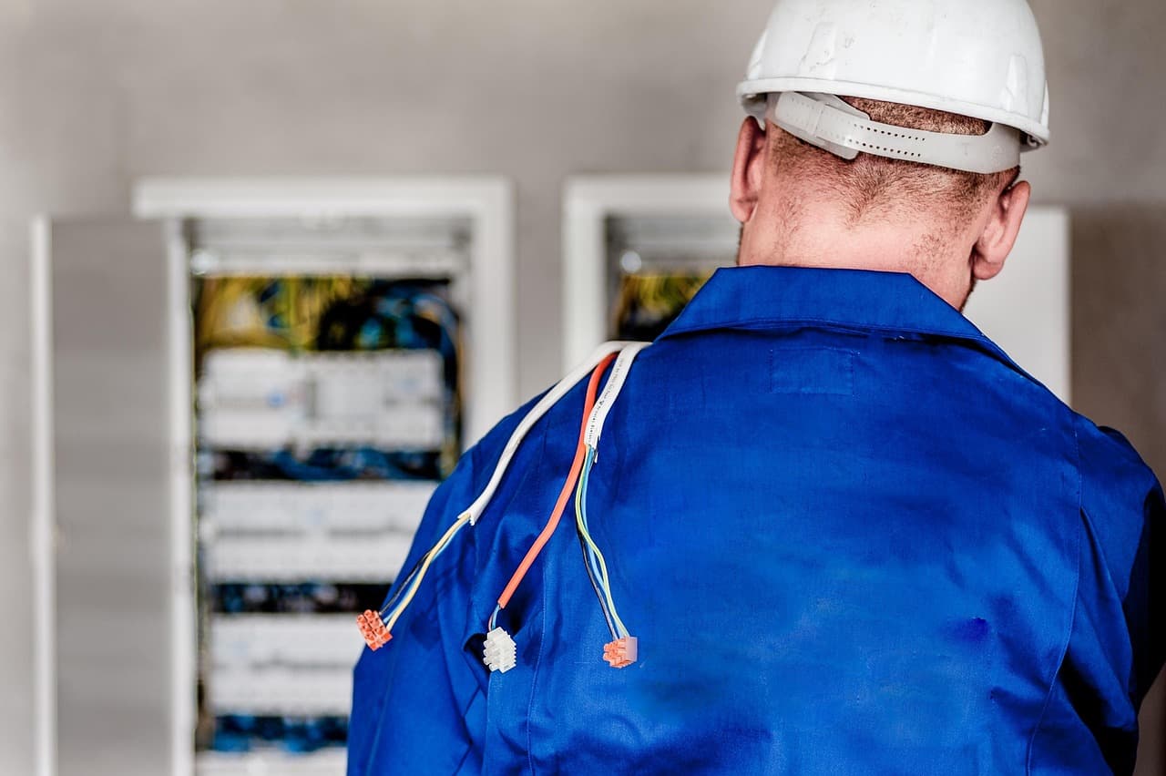 Mavi iş kıyafetli, baretli teknisyen elektrik panosunun önünde; arkadan